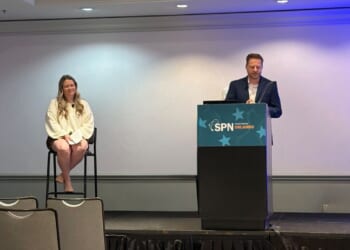 Camille Walsh, director of media relations at State Policy Network, sits on a tall chair, and Corey Burres, senior director of communication strategies at State Policy Network, stands at a podium labeled SPN Orlando in a conference room, with empty chairs in the foreground.