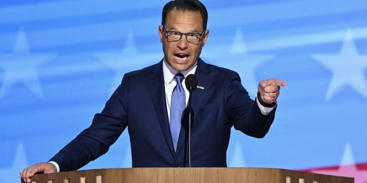 A man in a blue suit and tie speaks passionately at a wooden podium, pointing with his right hand. The background features a blue pattern with white stars, resembling the American flag.
