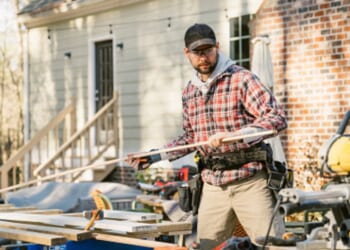 A man works in the yard of a house with wood and tools.
