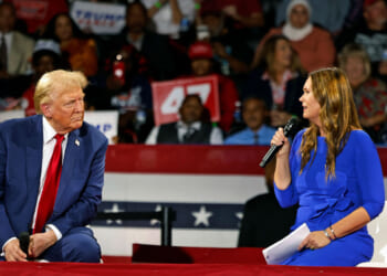 Donald Trump in blue suit and red tie sits on stage beside Sarah Huckabee Sanders in a blue dress holding a microphone, with several women visible among the crowd and patriotic banners in the background.