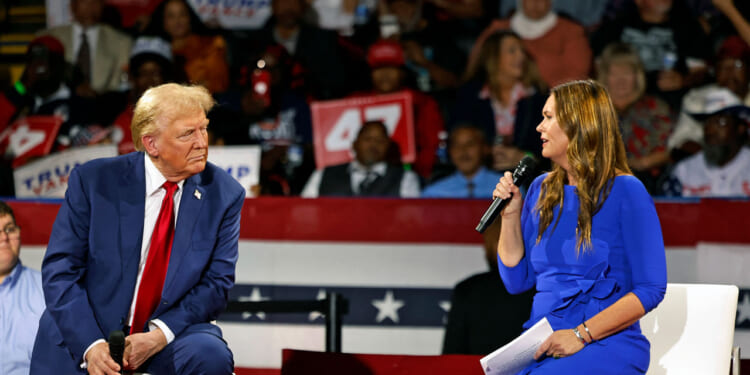 Donald Trump in blue suit and red tie sits on stage beside Sarah Huckabee Sanders in a blue dress holding a microphone, with several women visible among the crowd and patriotic banners in the background.