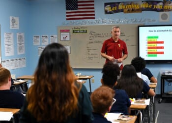 A teacher stands at the front of a classroom, speaking to students seated at desks. A large screen displays instructions, and educational posters are visible on the walls. An American flag hangs in the background.