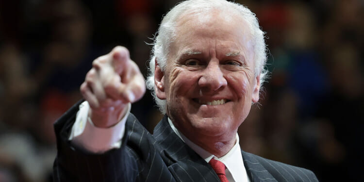 An older man in a suit and red tie smiles while pointing toward the camera. The background is blurred, focusing attention on his expression and gesture.