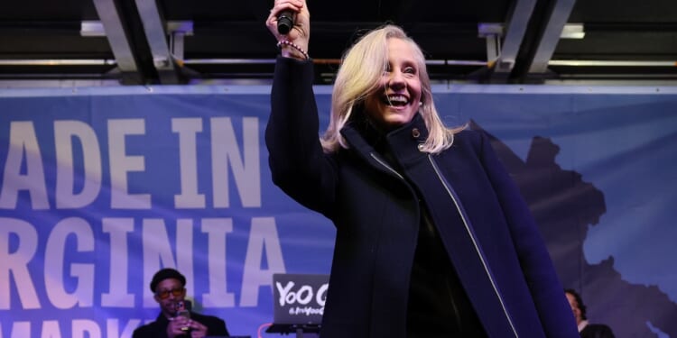 Abigail Spanberger with blonde hair smiles and raises a microphone on stage. A musician performs on equipment behind her. The background features a blue banner with partially visible text.