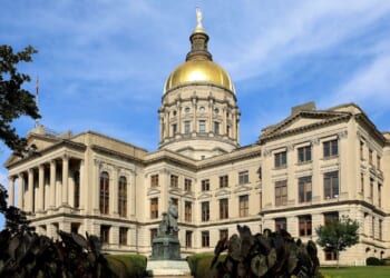 The Georgia State Capitol building with a prominent gold dome under a blue sky, surrounded by greenery and a statue in front.