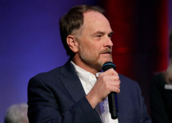 A man with short hair and a beard, wearing a blue blazer and white shirt, speaks into a microphone against a blue and red background, addressing the topic of Steve Toth Dyslexia.
