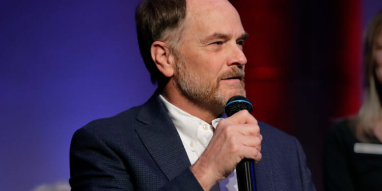 A man with short hair and a beard, wearing a blue blazer and white shirt, speaks into a microphone against a blue and red background, addressing the topic of Steve Toth Dyslexia.