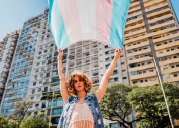 A trans person waves a transgender flag near a large building.