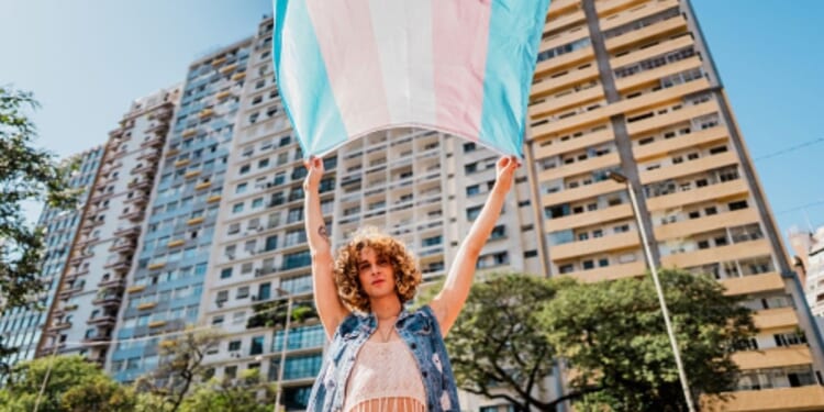 A trans person waves a transgender flag near a large building.