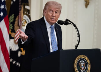A man in a suit speaks at a podium with the presidential seal, gesturing with one hand. U.S. flags and a decorated curtain are visible in the background.