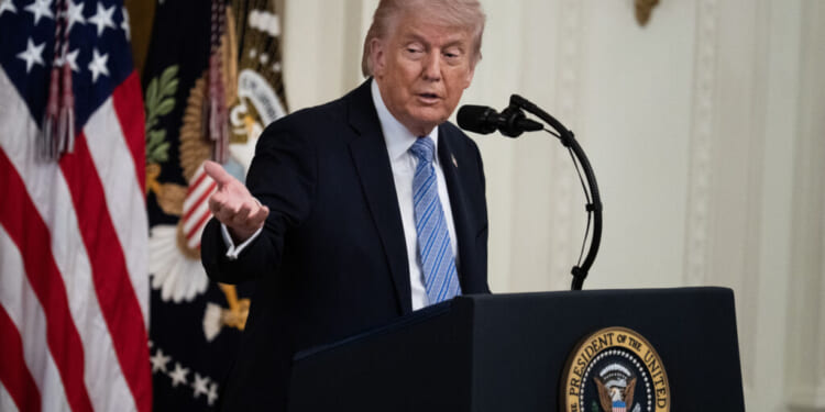 A man in a suit speaks at a podium with the presidential seal, gesturing with one hand. U.S. flags and a decorated curtain are visible in the background.