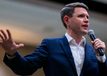 James Talarico in a navy suit and white shirt speaks into a microphone, gesturing with one hand, in a brightly lit indoor setting.