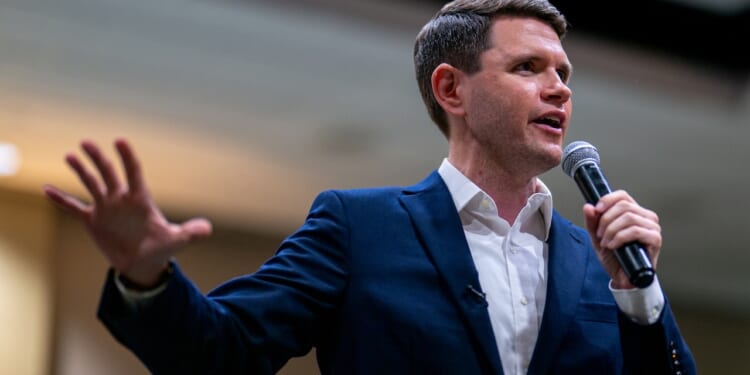 James Talarico in a navy suit and white shirt speaks into a microphone, gesturing with one hand, in a brightly lit indoor setting.