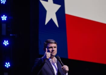A man in a suit gives a thumbs up while holding a microphone, standing in front of a large Texas flag projected on a screen. Blue stage lights are visible on the left.