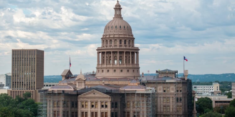 The Texas State Capitol building in Austin, with scaffolding on parts of its exterior, stands under a cloudy sky. The Texas flag is visible on the right side.