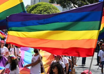 People march in a parade holding rainbow pride flags. Many wear white shirts and colorful accessories, celebrating LGBTQ+ pride outdoors under trees and sunlight.