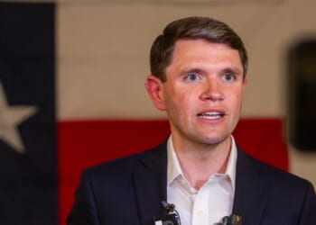 A man in a suit speaks in front of a large Texas flag backdrop, with microphones clipped to his jacket.