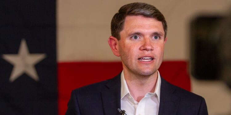 A man in a suit speaks in front of a large Texas flag backdrop, with microphones clipped to his jacket.
