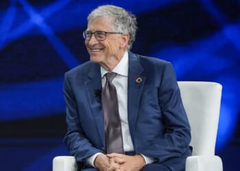 An older man in a blue suit and glasses sits smiling on a white chair against a blue background at a formal event.