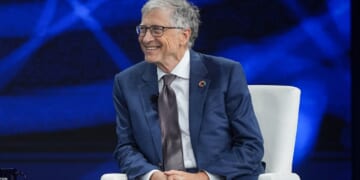 An older man in a blue suit and glasses sits smiling on a white chair against a blue background at a formal event.