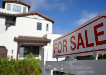 A white house with a brown roof is pictured behind a large For Sale sign in the front yard, under a blue sky with scattered clouds.