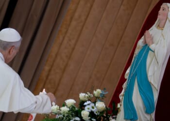 A person in white robes lights a candle beside a bouquet of flowers and a statue of the Virgin Mary, who is dressed in white and blue and has hands together in prayer.