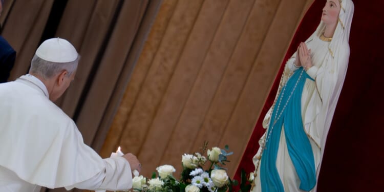 A person in white robes lights a candle beside a bouquet of flowers and a statue of the Virgin Mary, who is dressed in white and blue and has hands together in prayer.