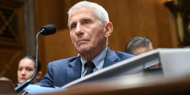 Dr. Anthony Fauci in a suit sits at a desk with a microphone, appearing serious, in a formal wooden room. Other people are seated in the background.