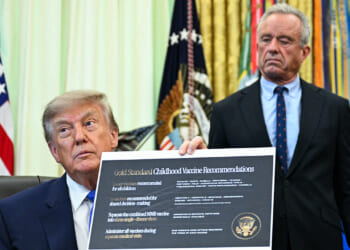 A man in a suit holds up a document titled Gold Standard Childhood Vaccine Recommendations while another man stands behind him in an official-looking room with flags in the background.