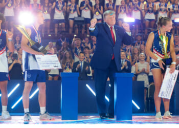 President Donald Trump dances on stage with young athletes holding trophies and checks at the Patriot Games. A cheering audience is in the background, making the scene appear festive and celebratory.