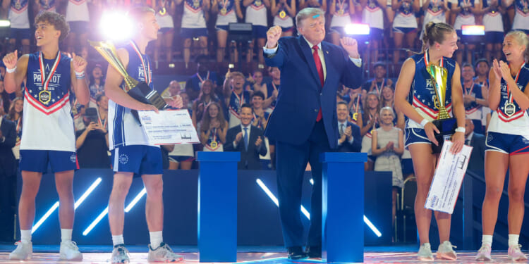 President Donald Trump dances on stage with young athletes holding trophies and checks at the Patriot Games. A cheering audience is in the background, making the scene appear festive and celebratory.