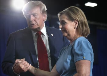 A man in a suit and red tie holds hands with a woman in a blue dress. They appear to be at an event, with blue curtains and bright stage lights in the background.