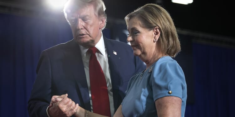 A man in a suit and red tie holds hands with a woman in a blue dress. They appear to be at an event, with blue curtains and bright stage lights in the background.