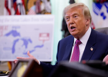 A man in a suit speaks while seated at a desk, gesturing with his hands. A map of the Great Lakes is visible in the blurred background, along with flags and ornate decor.