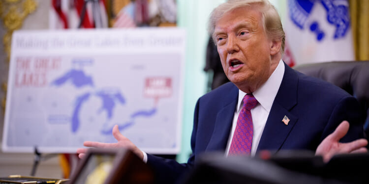A man in a suit speaks while seated at a desk, gesturing with his hands. A map of the Great Lakes is visible in the blurred background, along with flags and ornate decor.