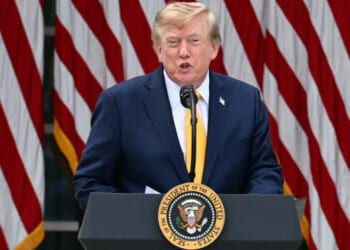 A man in a suit speaks at a podium with the U.S. presidential seal, with several large American flags in the background.