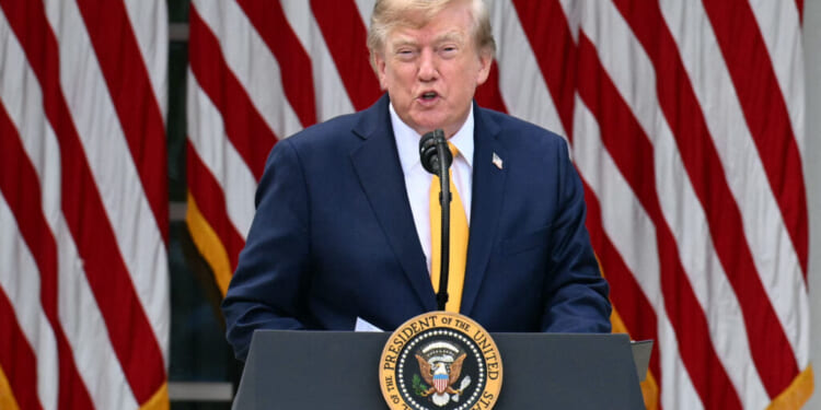 A man in a suit speaks at a podium with the U.S. presidential seal, with several large American flags in the background.