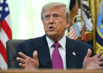 President Donald Trump in a suit sits at a desk, gesturing with his hands while speaking. U.S. flags and a presidential seal are visible in the background.