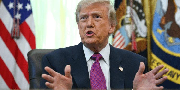 President Donald Trump in a suit sits at a desk, gesturing with his hands while speaking. U.S. flags and a presidential seal are visible in the background.