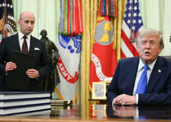 A man in a suit stands holding a folder while another man sits at a large desk, both in a room with military flags, the U.S. flag, and a gold curtain in the background.