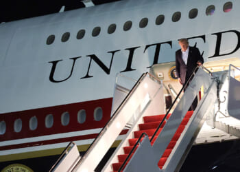A man descends the stairs from an airplane with UNITED written on its side. The steps are red-carpeted, and it is nighttime. The presidential seal is visible near the entrance.