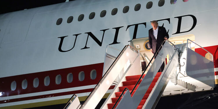 A man descends the stairs from an airplane with UNITED written on its side. The steps are red-carpeted, and it is nighttime. The presidential seal is visible near the entrance.