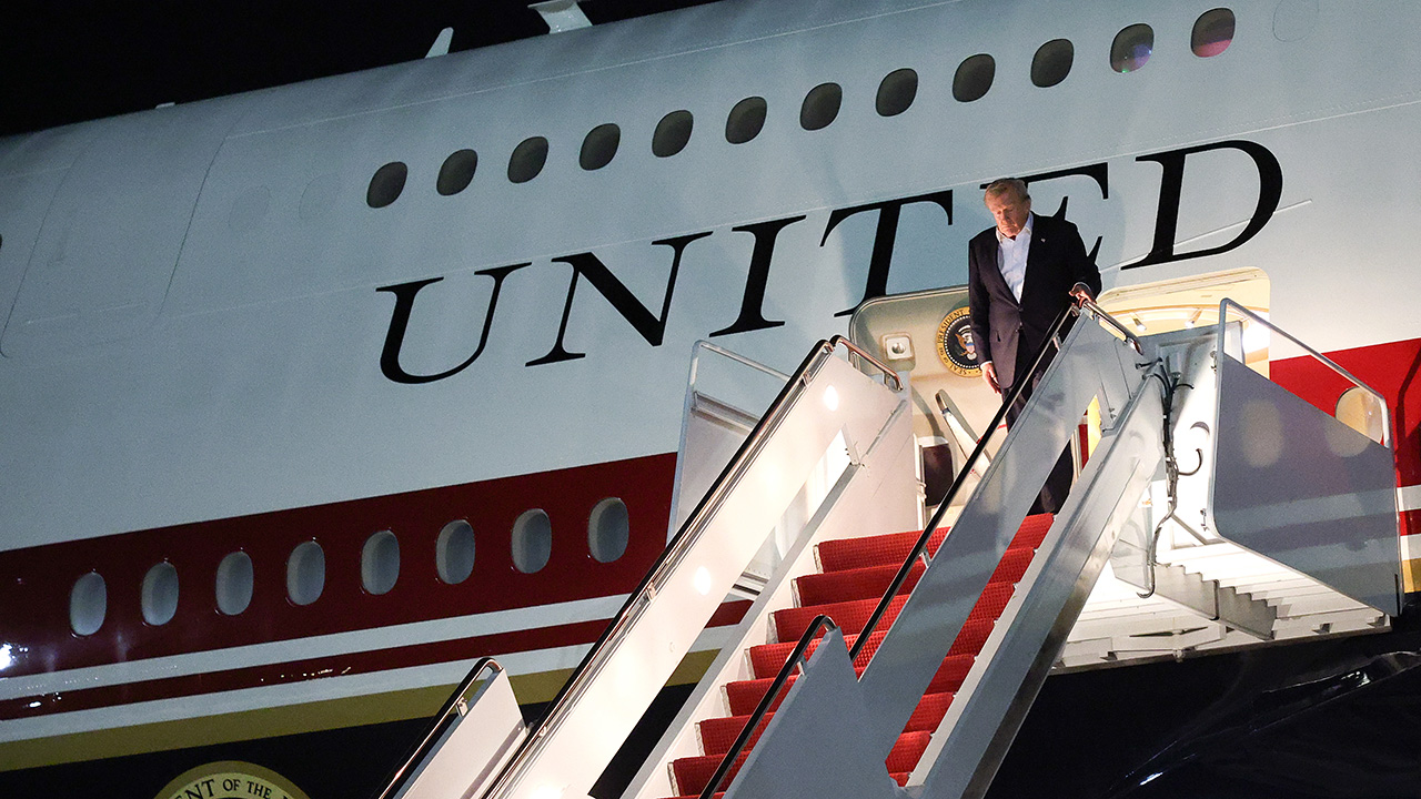A man descends the stairs from an airplane with UNITED written on its side. The steps are red-carpeted, and it is nighttime. The presidential seal is visible near the entrance.