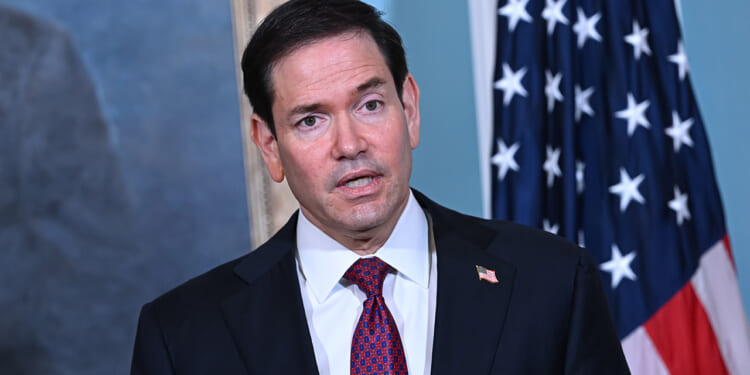 Marco Rubio in a suit and red patterned tie speaks in front of a U.S. flag and a painting, with an American flag pin on his lapel.