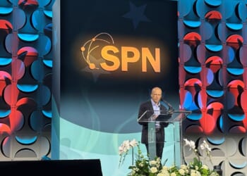 Speaker Yuval Levin stands at a podium on a decorated stage with the letters SPN prominently displayed behind him. Flowers are arranged in front of the podium, and the background features a geometric pattern with blue and red lights.
