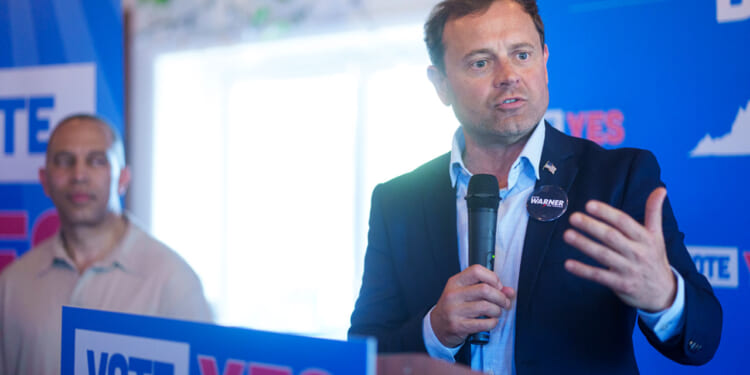 Tom Perriello speaks into a microphone at a podium with VOTE YES signs, while Hakeem Jeffries stands in the background.