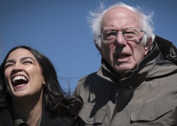 Two people stand outdoors against a blue sky; one is laughing with mouth wide open, the other, older, with white hair and glasses, looks forward with a neutral expression, both wearing jackets.