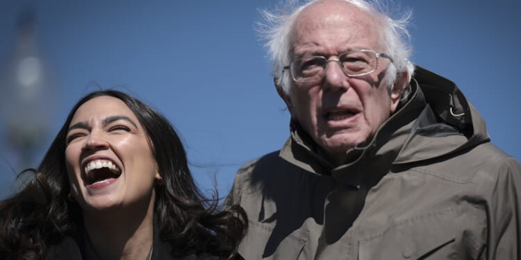 Two people stand outdoors against a blue sky; one is laughing with mouth wide open, the other, older, with white hair and glasses, looks forward with a neutral expression, both wearing jackets.