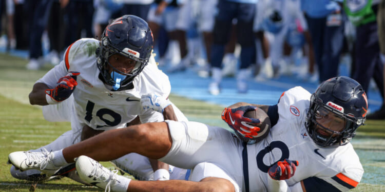 Two football players in white Virginia uniforms are on the field; one is holding the ball while laying down, and the other is behind him, with a blurred crowd and players in blue in the background.