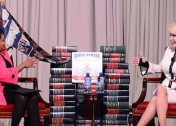 Dolly parton and another woman sit facing each other, surrounded by stacks of books. One wears a pink jacket and gestures with a scarf, while the other, in a white outfit, smiles. A book titled Dolly Parton is displayed between them.